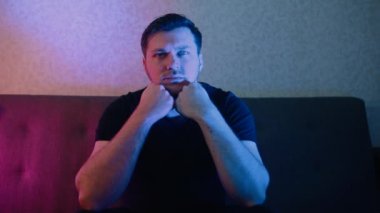 Kanepede oturan bir adam dikkatle televizyon izler, ellerine yaslanır..