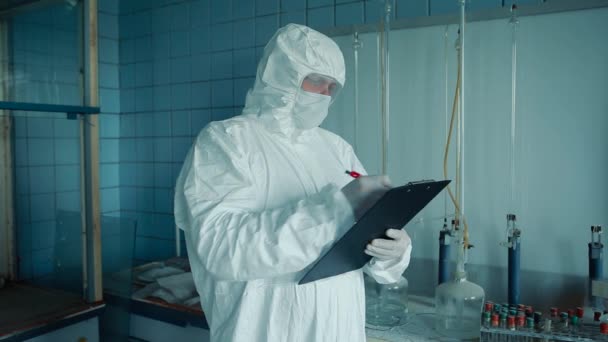 Pracownik medyczny w kombinezonie ochronnym i okularach posiada teczkę z dokumentami w rękach laboratorium w tle, przedstawiającymi dane. — Wideo stockowe