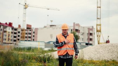Turuncu yelekli ve turuncu renkli bir adam kalbine yapışıp bir inşaat alanının arka planına ulaşmaya çalışıyor..