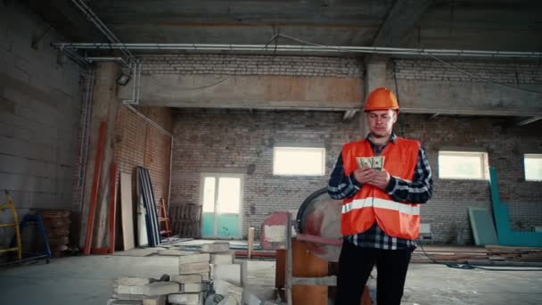 Een mannelijke bouwer staat in een gebouw te midden van een bouwplaats die geld vertelt.. — Stockvideo