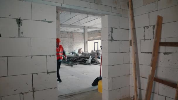 Un constructor masculino con chaleco naranja y casco naranja entra en la puerta, llevando consigo una gran pala . — Vídeos de Stock