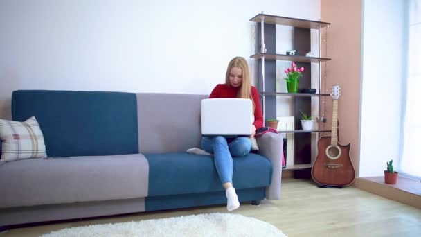 Een jonge vrouw in een rode trui zit op een bank en typt op afstand tekst achter een laptop. — Stockvideo