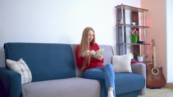 En flicka i en röd tröja sitter på en soffa sprider pengar fantiserar om vilket köp hon kommer att spendera. — Stockvideo