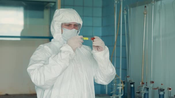 Un trabajador médico con un traje protector y gafas de seguridad sostiene un tubo de ensayo en sus manos . — Vídeos de Stock