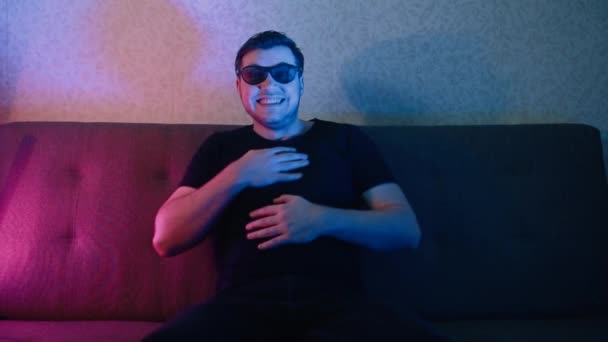 A young man sits on a sofa with 3D glasses and watches TV and is filled with emotions. — Stock Video