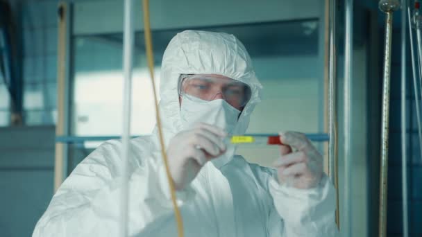 En läkare i skyddsdräkt och glasögon håller ett provrör i sina händer medan han undersöker det. — Stockvideo
