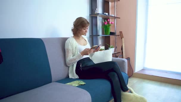 Uma menina insere dados de um cartão em um smartphone enquanto está sentada em um sofá . — Vídeo de Stock