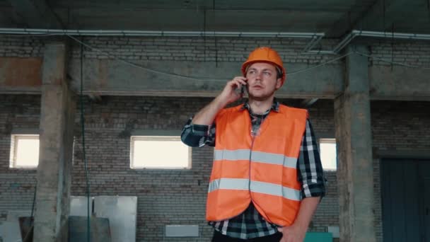 Construtor masculino em um colete laranja e capacete laranja no fundo de um canteiro de obras falando ao telefone . — Vídeo de Stock