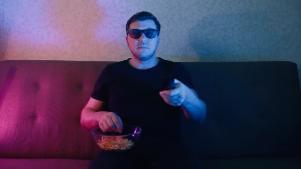 Un hombre se sienta en un sofá en gafas 3D comiendo patatas fritas y cambiando de canal en un televisor . — Vídeos de Stock