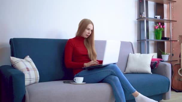 A young woman in a red sweater is sitting on a couch drinking coffee and working remotely pesatet text behind a laptop. — Stock Video