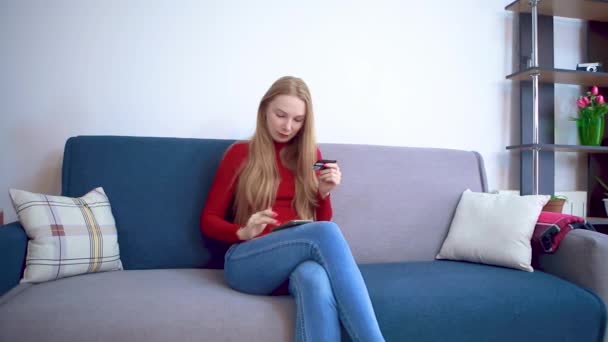 Een jong meisje in een rode trui op een bank voert gegevens van een creditcard in de telefoon. — Stockvideo