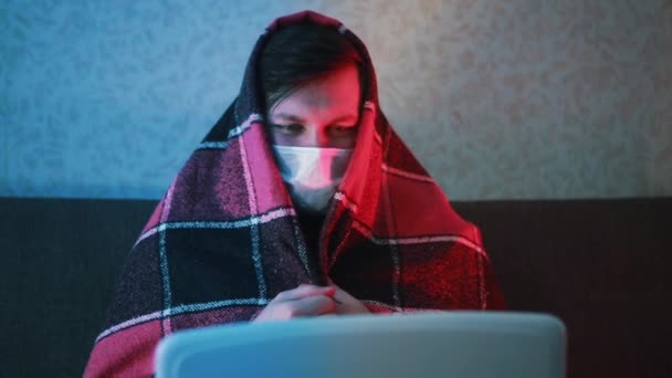 En man insvept i en filt och i en mask sitter i karantän och kommunicerar på en laptop. — Stockvideo