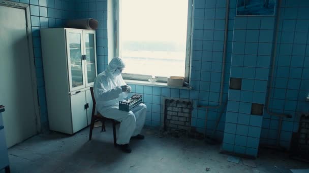 保護服とゴーグルを着た男が研究室の椅子に座って分析でフラスコを調べます. — ストック動画