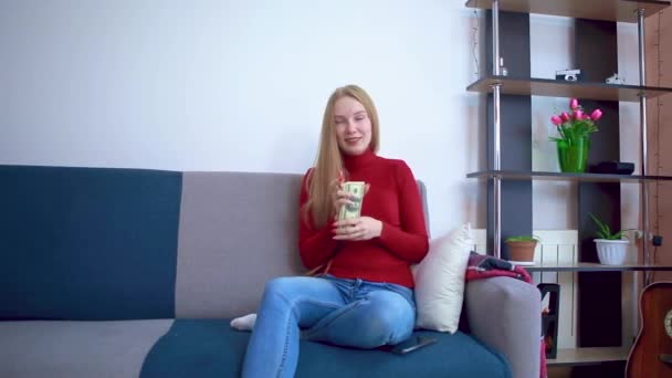 Dziewczyna w czerwonym swetrze siedząca na kanapie z radością liczy pieniądze i marzy o czymś dobrym.. — Wideo stockowe