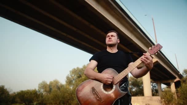 Een jongeman speelt een akoestische gitaar onder de brug. — Stockvideo