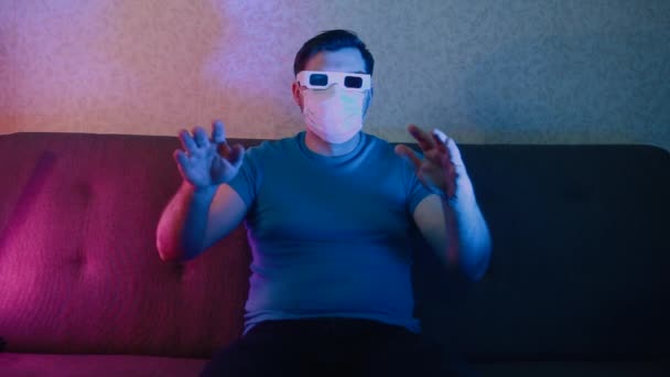Um homem com uma máscara protetora está sentado em um sofá usando óculos estéreo, assistindo TV acenando com as mãos . — Vídeo de Stock