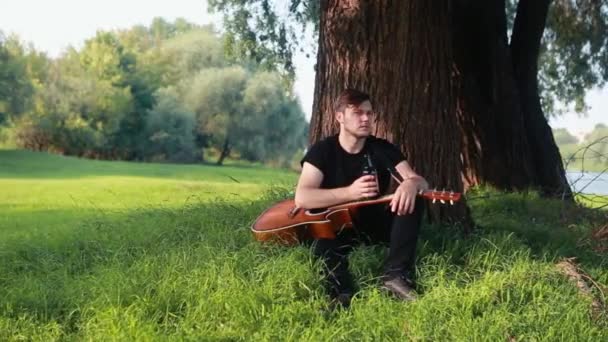 Молодой парень сидит под деревом в руках с гитарой и пьет бутылку пива. — стоковое видео