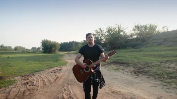 Un joven camina por un camino polvoriento y toca la guitarra . — Vídeos de Stock