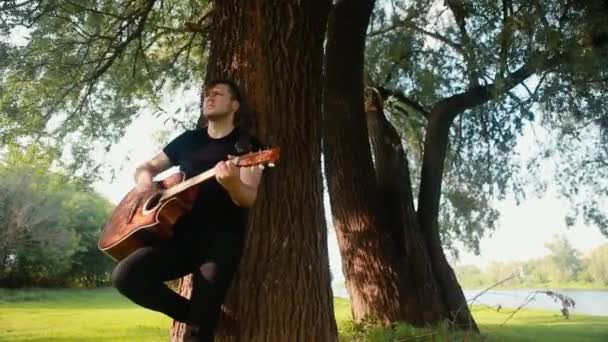 Młody mężczyzna w czarnej koszulce opierający się na drzewie trzyma w rękach gitarę akustyczną i na niej gra.. — Wideo stockowe