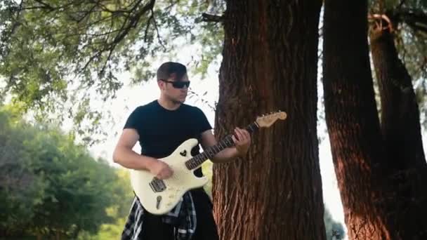 Een jongeman in een zwart T-shirt speelt een elektrische gitaar bij een boom. — Stockvideo