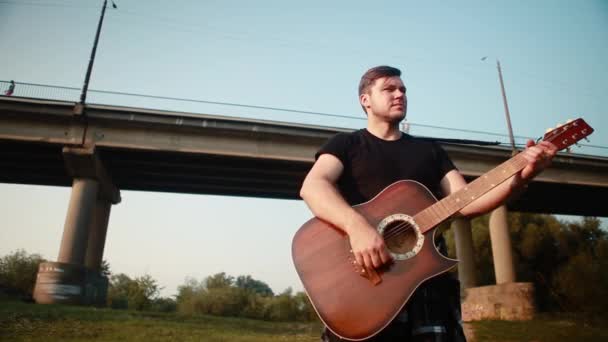 Młody mężczyzna w czarnej koszulce stoi na tle mostu i gra na gitarze akustycznej. — Wideo stockowe