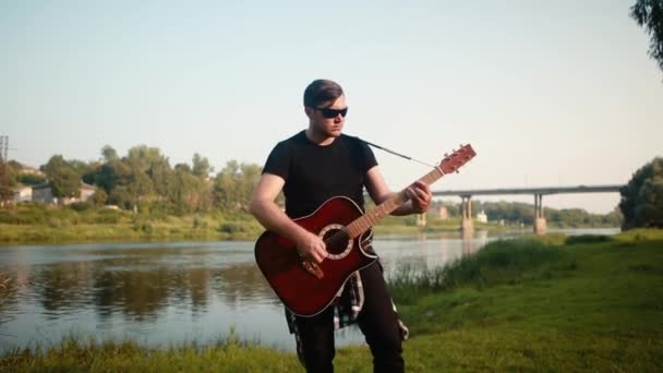 Um homem de t-shirt preta e óculos pretos toca uma guitarra acústica no fundo de uma ponte e de um rio . — Vídeo de Stock