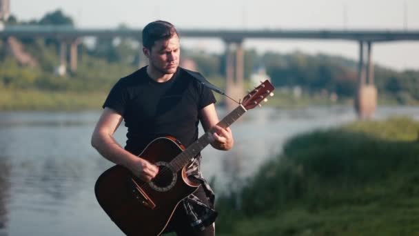 Un hombre con una camiseta negra rápidamente toca una guitarra acústica contra un puente y un río . — Vídeos de Stock