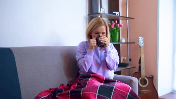 Ung blond kvinna som sitter på en soffa insvept i en filt dricker en drink från en mugg. — Stockvideo