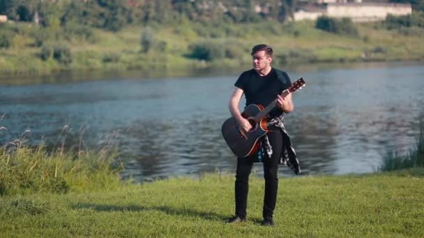 Een mannelijke gitarist speelt een akoestische gitaar tegen de achtergrond van een rivier. — Stockvideo