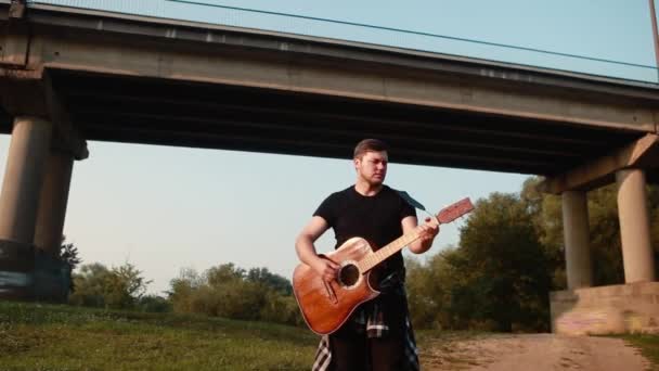 Młody facet gitarzysta gra na gitarze akustycznej pod mostem z przejeżdżającymi samochodami. — Wideo stockowe