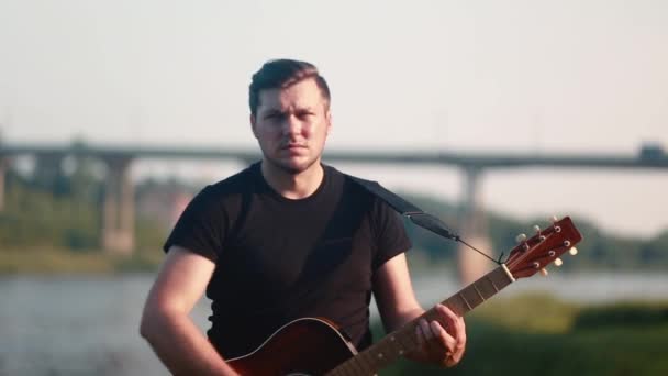 Ein männlicher Gitarrist spielt eine Akustikgitarre vor dem Hintergrund einer Brücke mit vorbeifahrenden Autos in Nahaufnahme. — Stockvideo