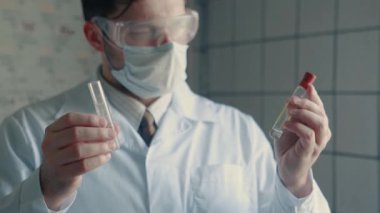 Koruyucu kıyafetli ve gözlüklü bir sağlık görevlisi elinde iki test tüpü tutuyor..