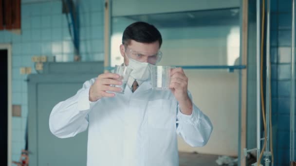 Um médico masculino de fato e máscara protetores segura tubos de ensaio nas mãos, derramando um concentrado entre eles. . — Vídeo de Stock