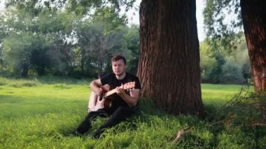Gitarist genç bir adam bir yaz parkında çimlerin üzerinde oturan bir ağacın yanında akustik gitar çalıyor..