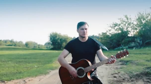 A male guitarist is walking along the road and playing an acoustic guitar. — Stock Video