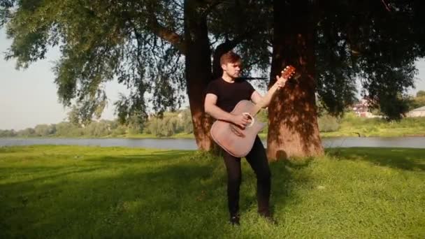 En ung kille en gitarrist spelar en akustisk gitarr nära ett träd i en sommarpark på bakgrunden av floden. — Stockvideo