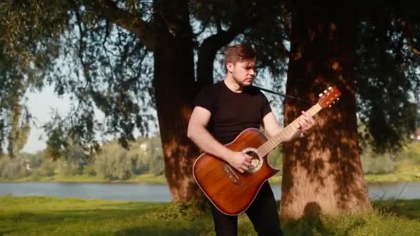 Ένας νεαρός με μαύρο μπλουζάκι παίζει ακουστική κιθάρα σε φόντο δέντρου και ποταμού.. — Αρχείο Βίντεο