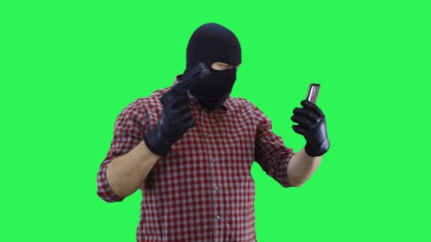 A man in a balaclava in a plaid shirt is talking on the phone with an interlocutor, threatening him with a gun. — Stock Video