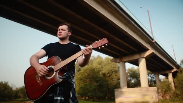 Un guitarrista con una camiseta negra toca una guitarra acústica bajo un puente . — Vídeos de Stock