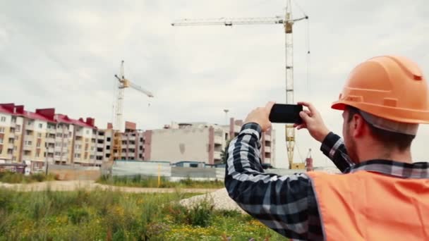 Muž v oranžové vestě a oranžové helmě natáčí stavební video na mobilním telefonu. — Stock video