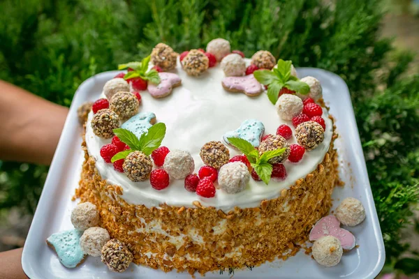 Really Handmade cake with cream, candys, leaves, hearts, coconuts. Cake in hand on a green background with wild berries and sweets.