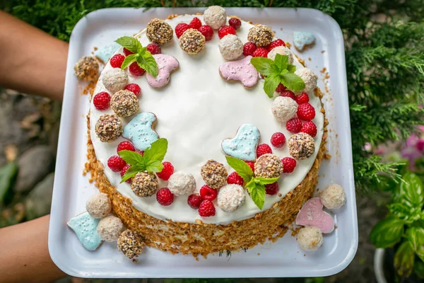 Really Handmade cake with cream, candys, leaves, hearts, coconuts. Cake in hand on a green background with wild berries and sweets.