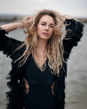 Güzel genç bir sarışın, portre, kameraya bakıyor, siyah bir mayo içinde, plaj peleriniyle, azgın bir denizin arka planında, rüzgarda dalgalanan saçlar yumuşak bir odak noktası.