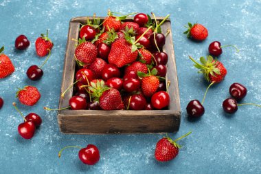 Taze olgun çilekler ve kirazlar ahşap kutuda mavi arka planda..