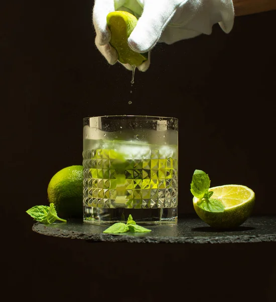 Donna che spreme succo di lime nel bicchiere di cristallo con rinfrescante Mojito. Cokctail di lime fresco con menta e cubetti di ghiaccio sul tavolo di pietra nera . — Foto Stock