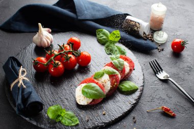 Mozzarella ile taş tabakta Caprese salatası, taze fesleğen yaprağı, domates, deniz tuzu, koyu arka planda kurutulmuş biber ve taze sarımsak. Deniz tuzu yiyeceğin üzerine düşer..