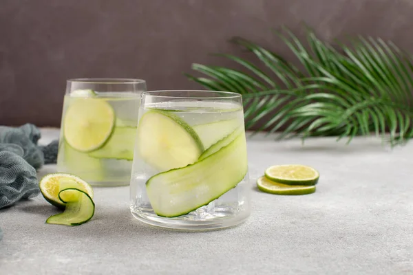 Acqua rinfrescante disintossicante con cetriolo fresco e fette di lime su tavolo di cemento grigio con foglie di palma.Cocktail di cetriolo sano. — Foto Stock