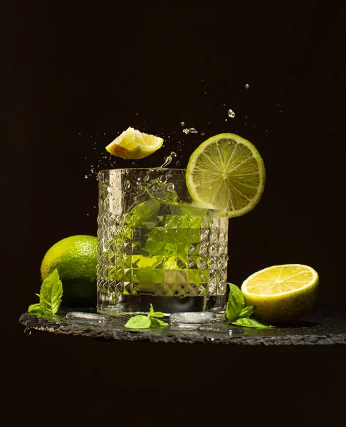 Bicchiere di cocktail Mojito con lime fresco, foglie di menta, cubetti di ghiaccio e acqua rinfrescante.Schizzo di cocktail su piatto di pietra nera . — Foto Stock