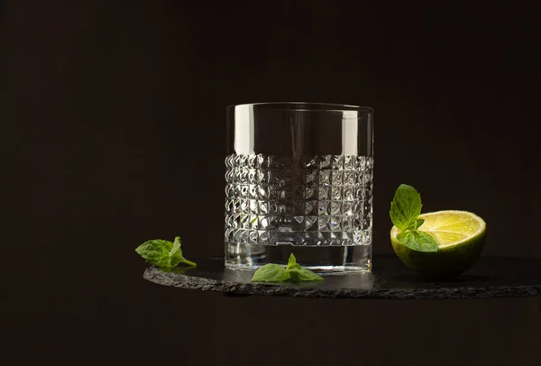 Bicchiere di cristallo vuoto su lastra di pietra nera con calce fresca e foglie di menta su sfondo scuro.. — Foto Stock
