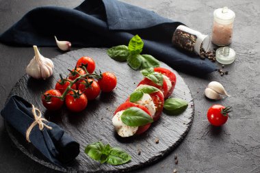 Mozzarella ile taş tabakta Caprese salatası, taze fesleğen yaprağı, domates, deniz tuzu, koyu arka planda kurutulmuş biber ve taze sarımsak. Deniz tuzu yiyeceğin üzerine düşer..